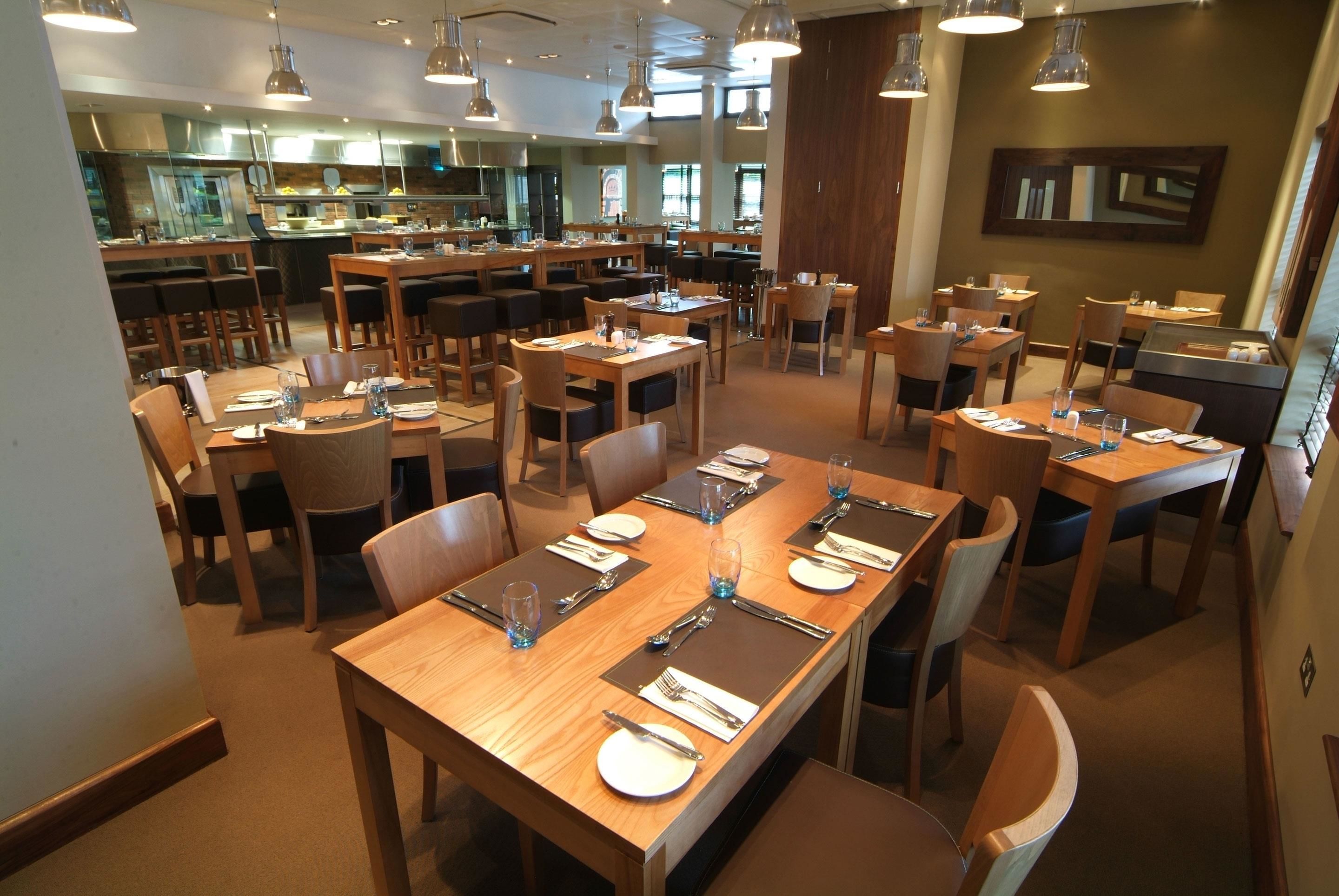 A spacious dining area with multiple tables and chairs, a kitchen in the background, and pendant lighting.