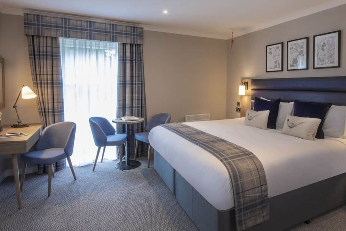 A hotel room with a large bed, a desk, two chairs, a small table, and a nightstand with a phone.