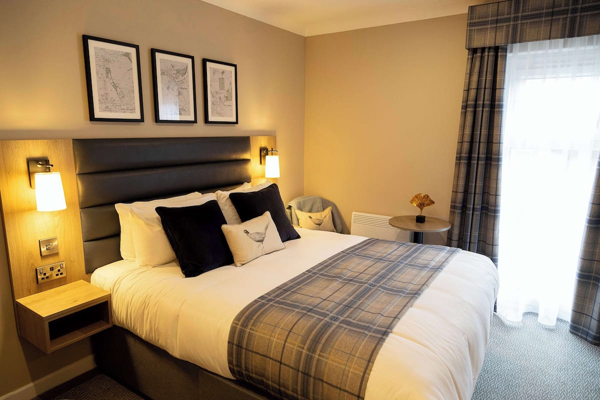 A hotel room with a large bed, two bedside tables with lamps, a small round table with a vase, and a window with curtains.