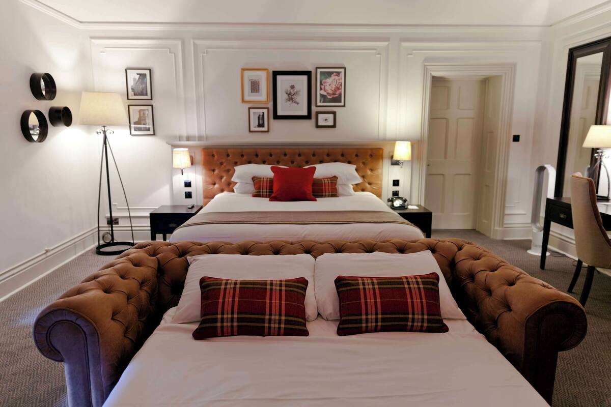A hotel room with a large bed, a smaller bed, a desk, and a chair. There are framed pictures on the wall and a floor lamp.