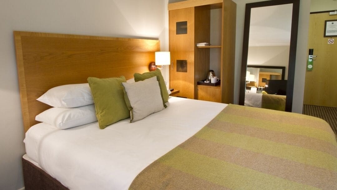Una habitació d'hotel amb un panell de melí a la capçal del llit, un llit amb roba de llit blanca, almoines verdes i blanques, una manta a ratlles, una tauleta de nit amb una làmpada i un armari amb un mirall.