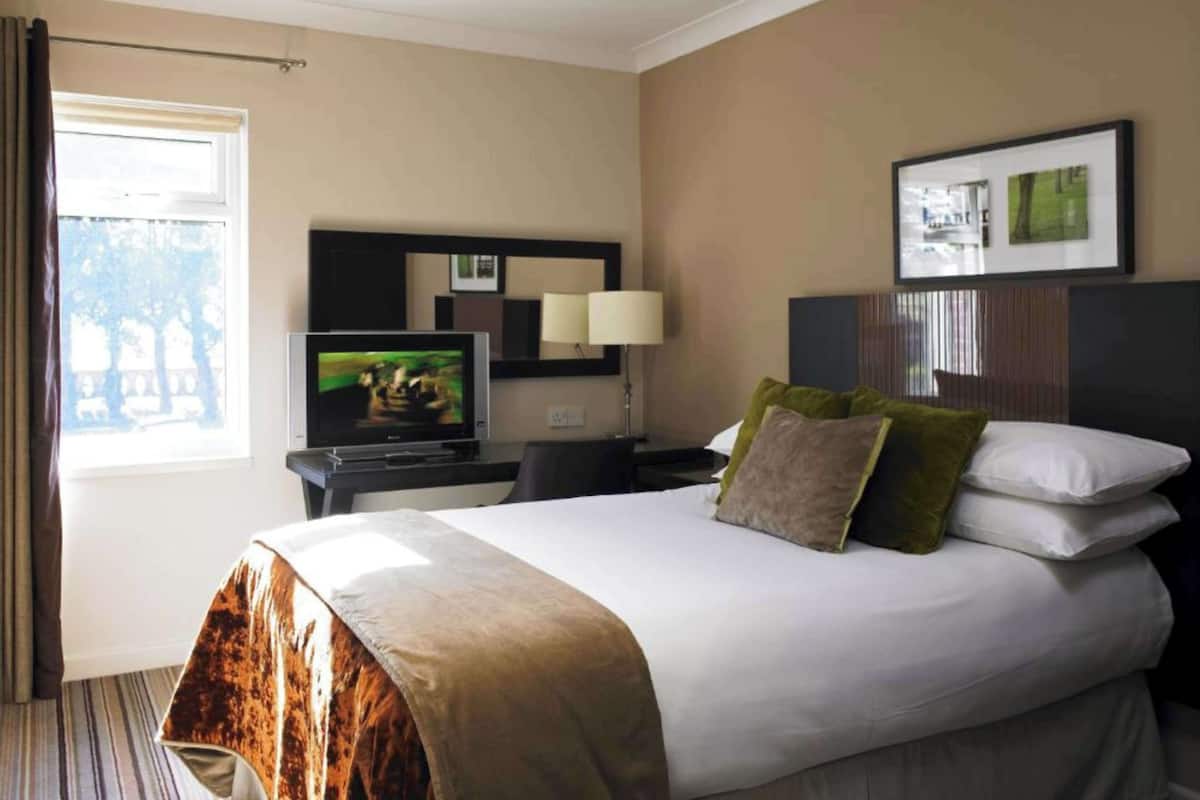 A hotel room with a bed, a TV, a nightstand, a lamp, and a window with curtains.