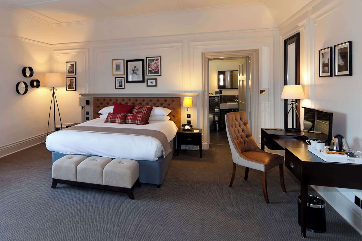A hotel room with a large bed, a desk, and a chair. There are framed pictures on the wall, a lamp, and a mirror.