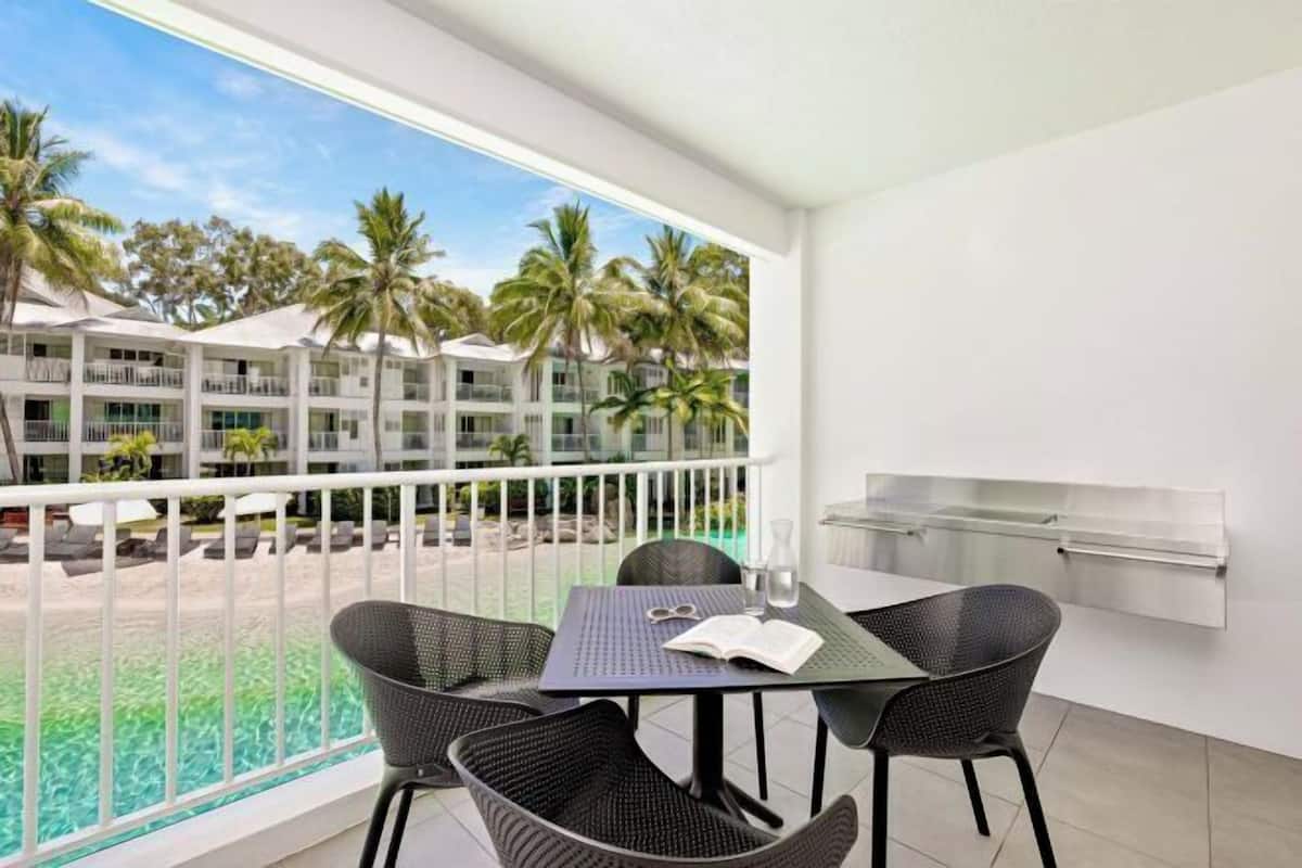 A balcony with a table and chairs, a grill, and a view of a pool and resort buildings.