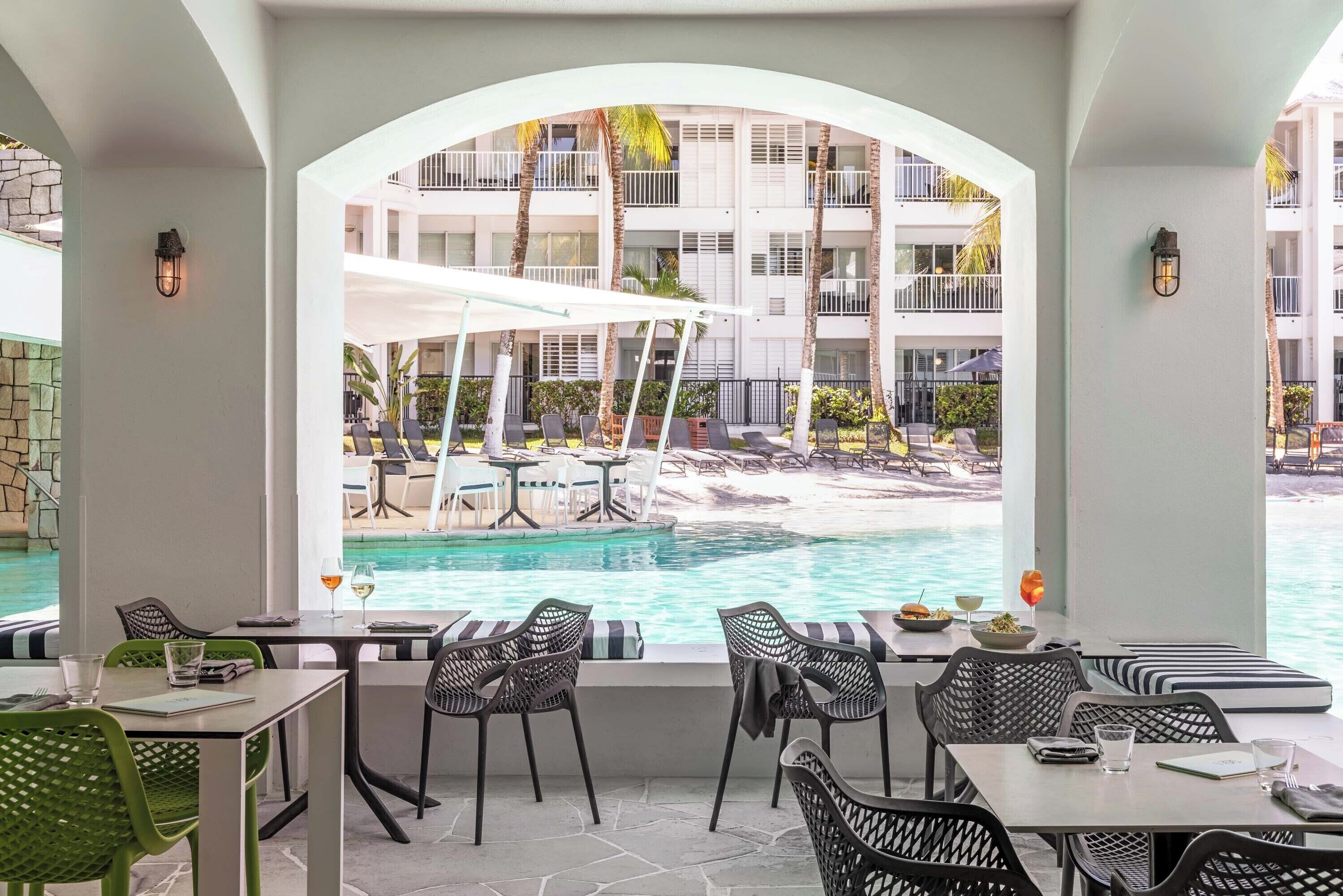 A poolside dining area with tables and chairs, a view of a pool, and a building with balconies in the background.