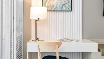 A modern hotel room with a white desk, a blue chair, a floor lamp, and a large abstract painting on the wall.