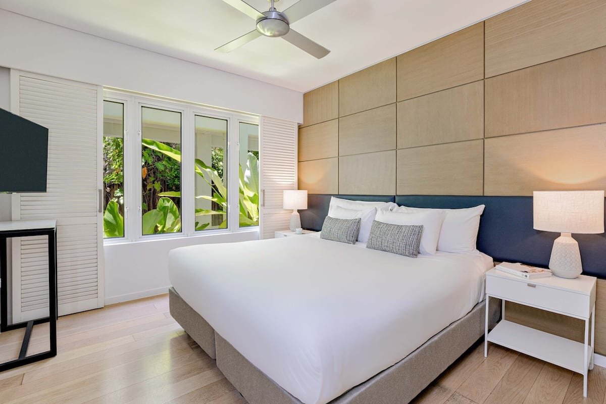 A modern bedroom with a large bed, a ceiling fan, a nightstand, and a window with a view of greenery.