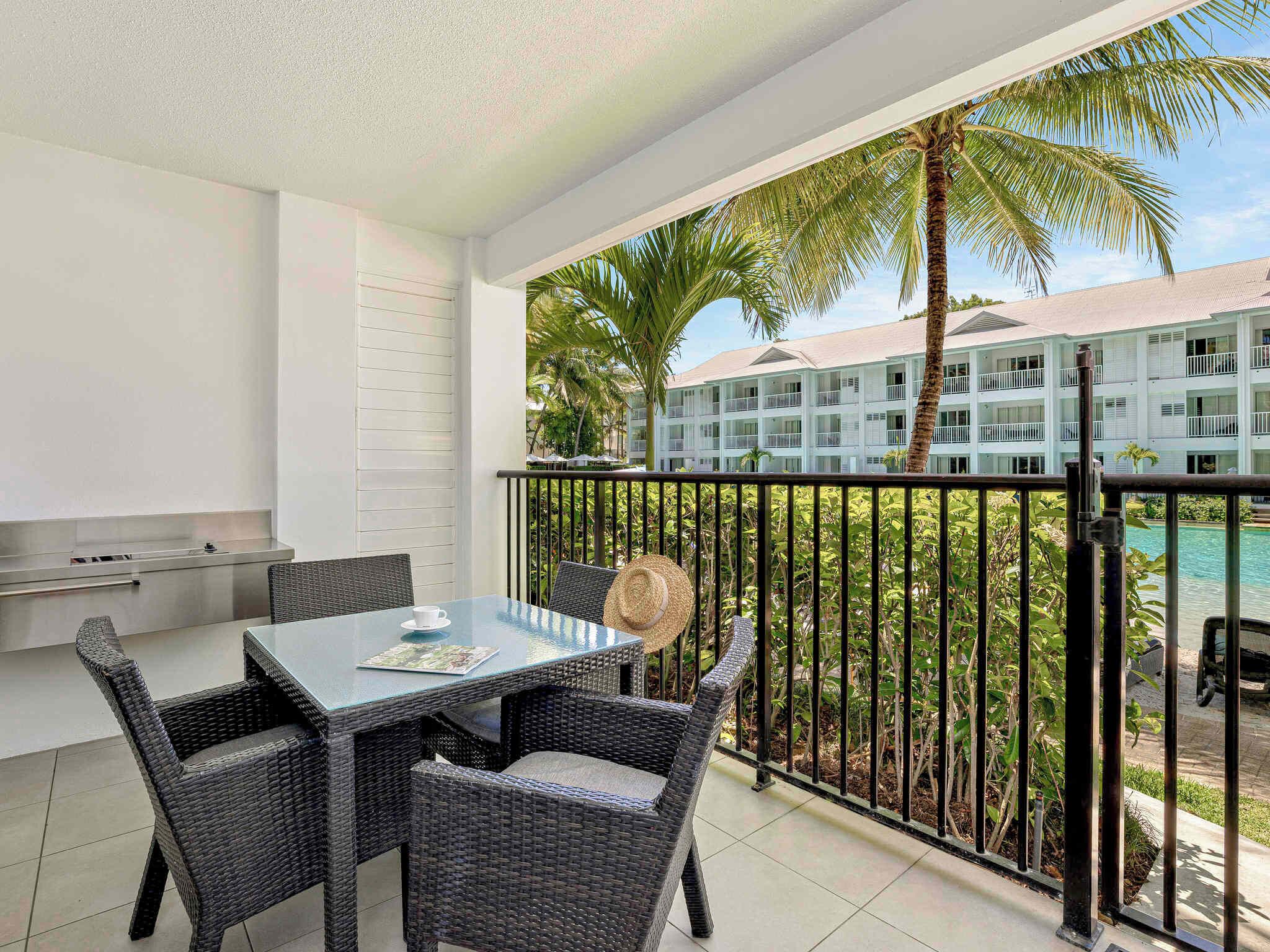 A balcony with a table and chairs, a grill, and a view of a pool and hotel building.