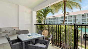 A balcony with a table and chairs, a grill, and a view of a pool and hotel building.