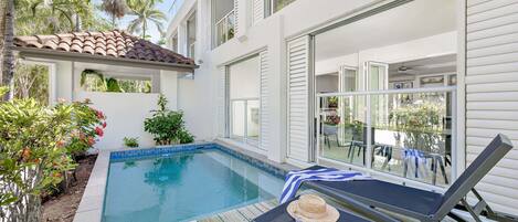 A swimming pool with a wooden deck, lounge chairs, and a view of a garden with palm trees.