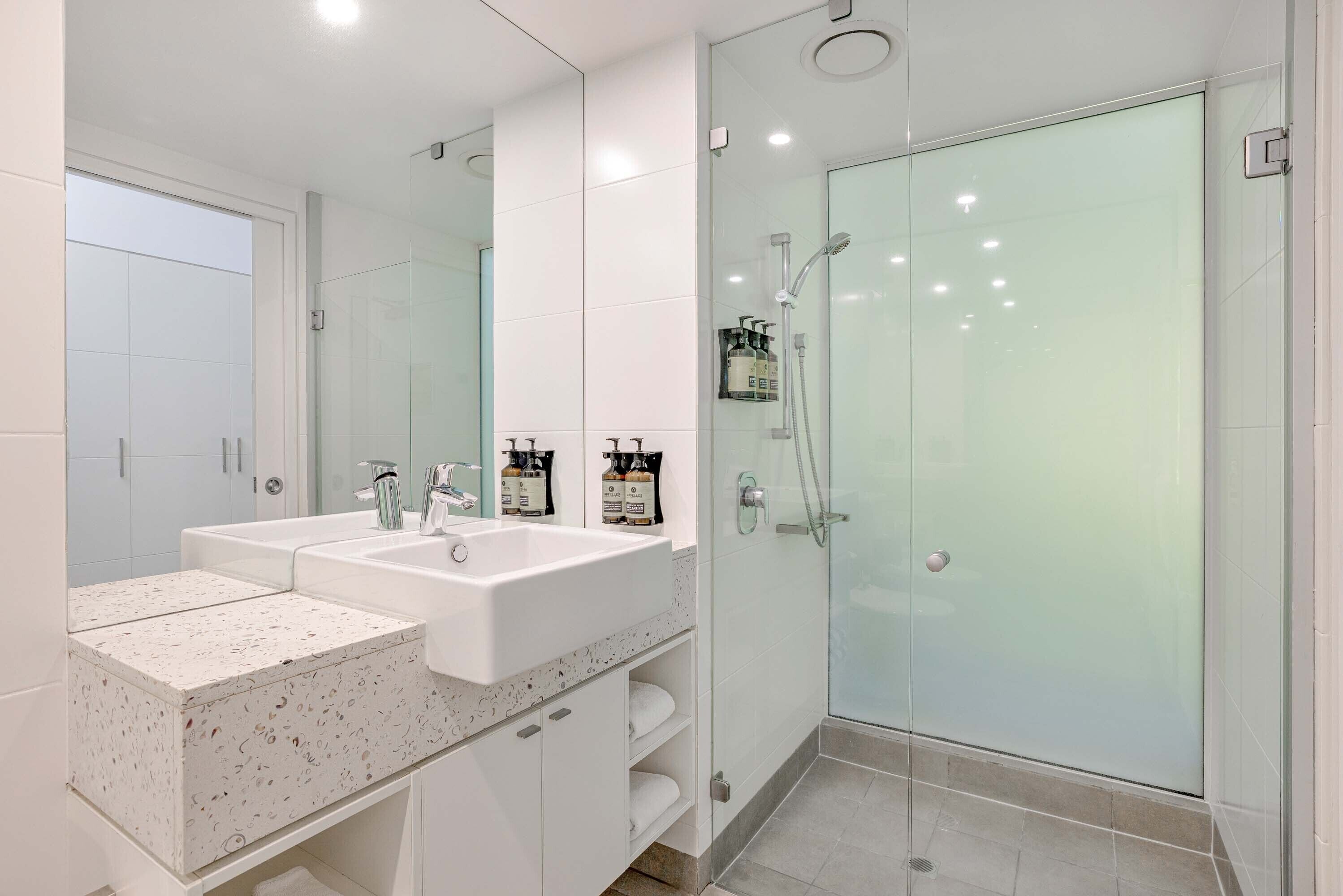 Un baño moderno con dos lavamanos, cabina de ducha con mampara de vidrio y un espejo grande.