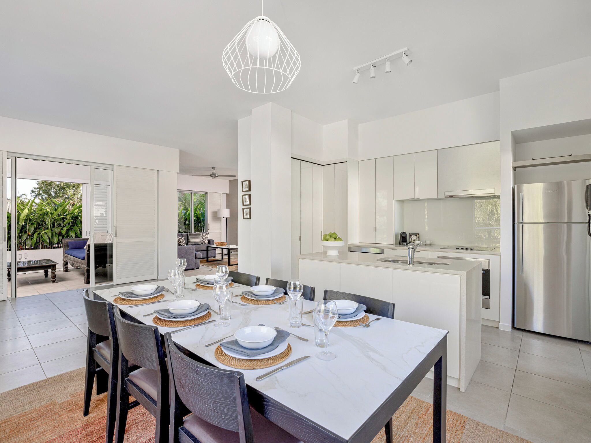 Une cuisine moderne dotée d’un îlot central, d’appareils électroménagers en acier inoxydable et d’un coin repas avec une table blanche dressée pour six personnes.