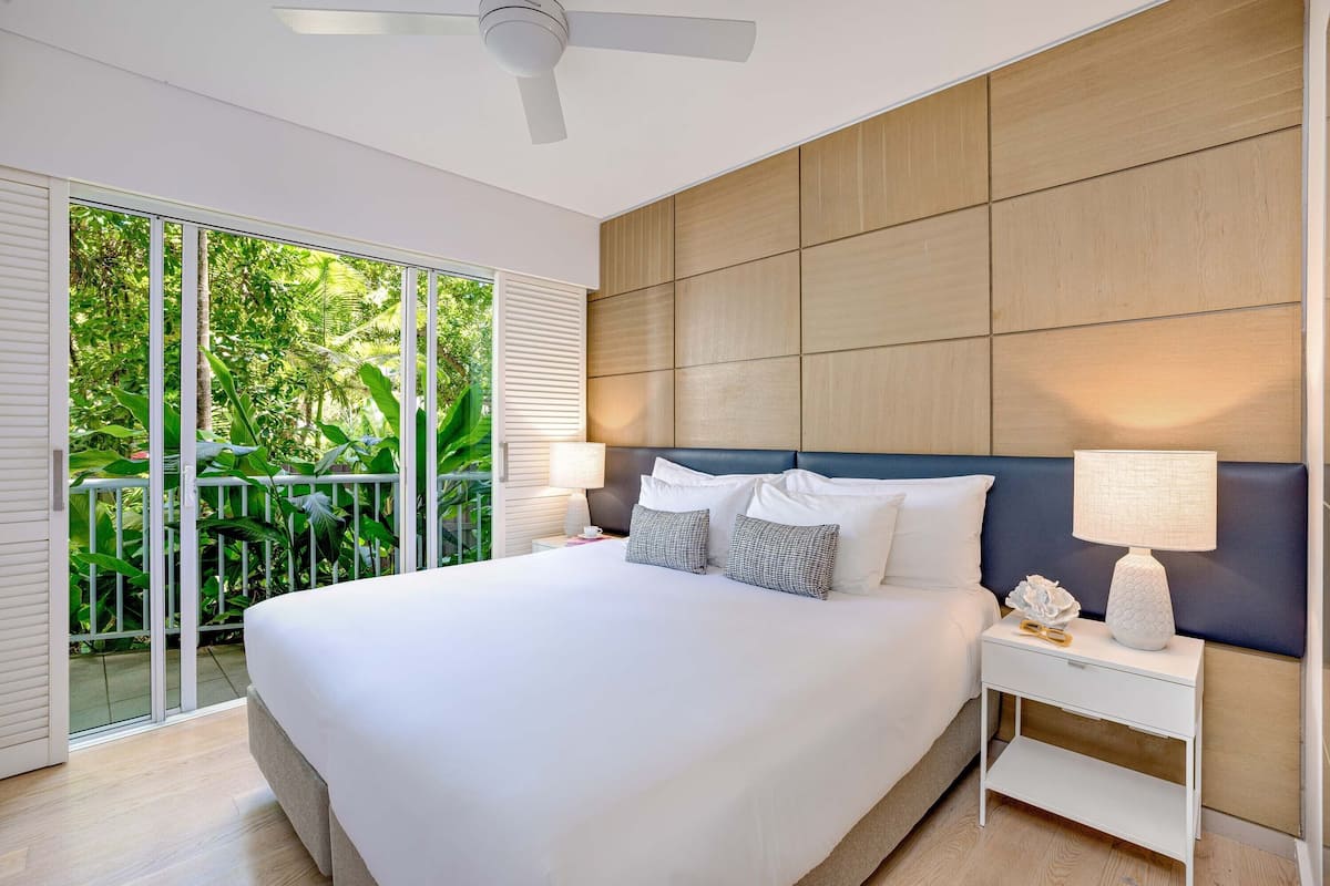 A modern bedroom with a large bed, a nightstand, a ceiling fan, and a view of lush greenery through a sliding glass door.