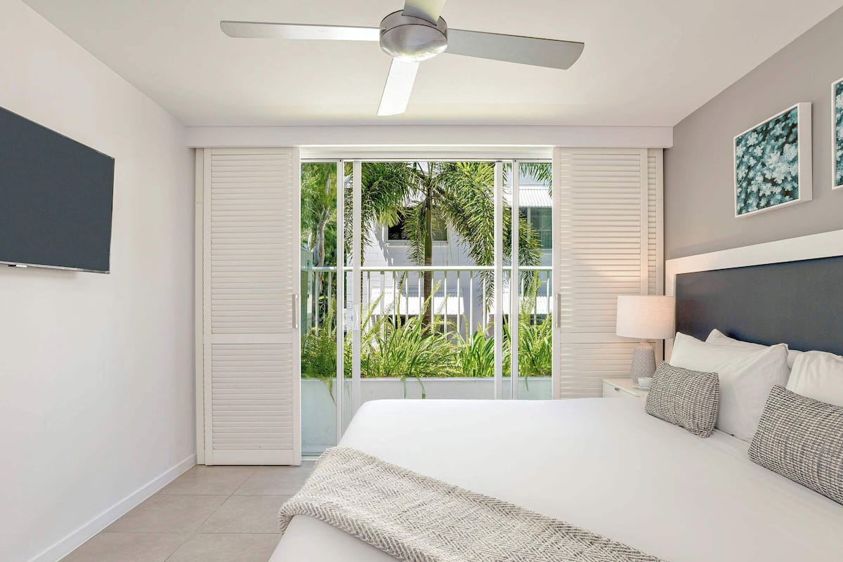 A modern bedroom with a large bed, a ceiling fan, and a view of greenery through a sliding door.