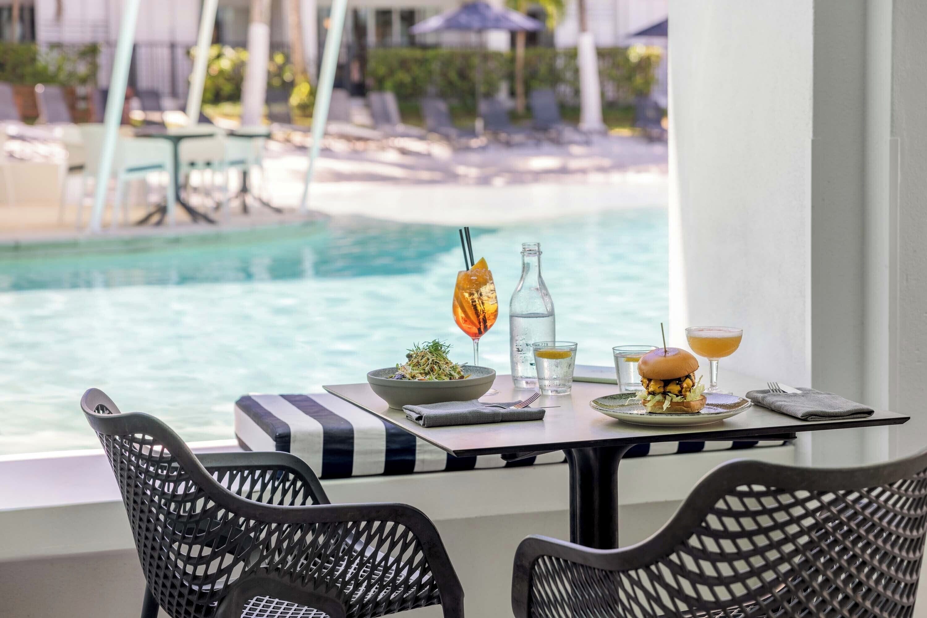 A dining table with a salad, burger, and drinks, overlooking a pool.