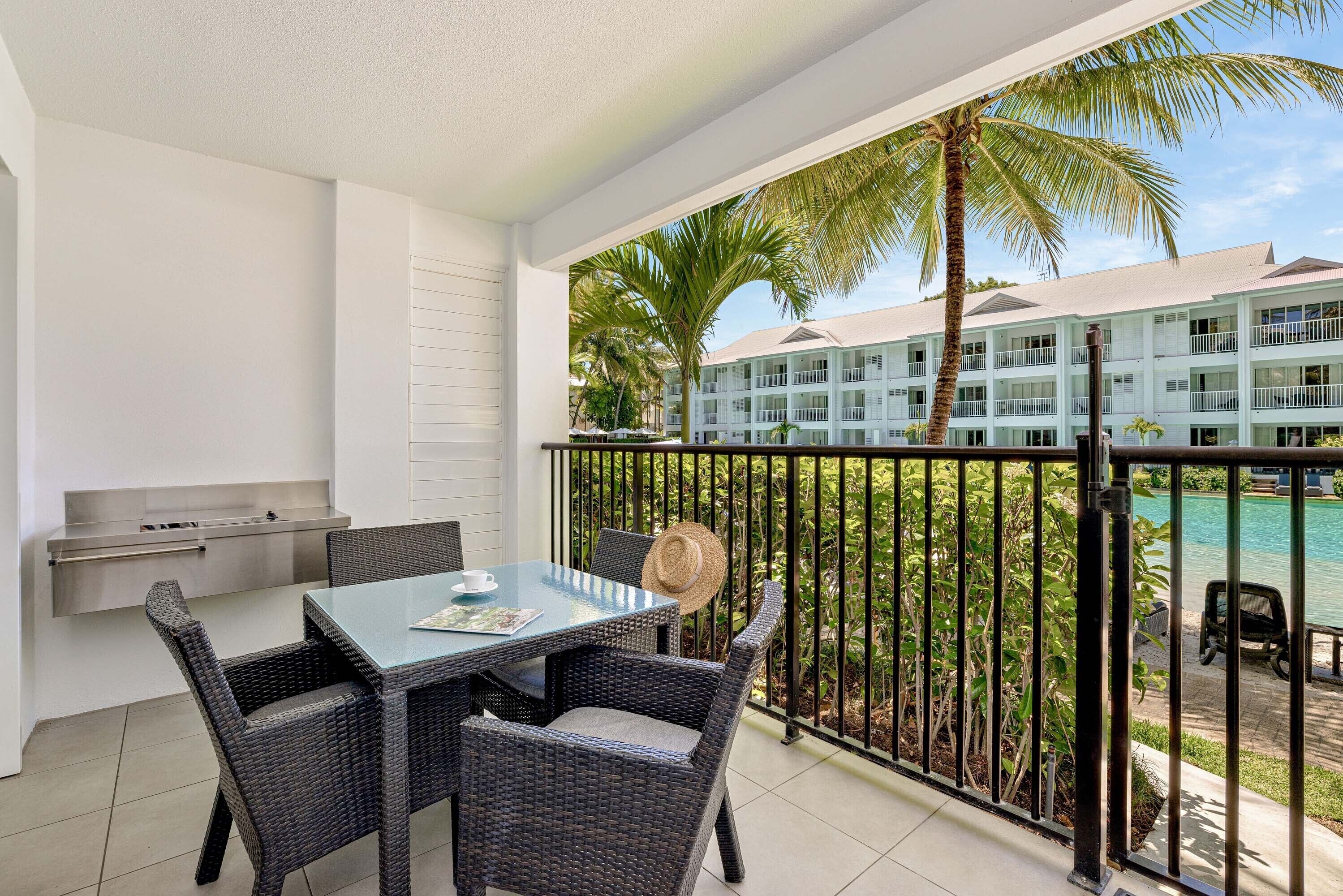 A balcony with wicker furniture, a glass table, and a view of a pool and hotel building.