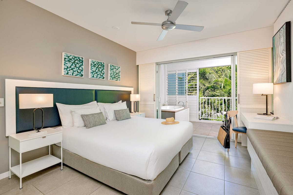 A modern hotel room with a large bed, a ceiling fan, a balcony with a view of greenery, and a desk with a chair.