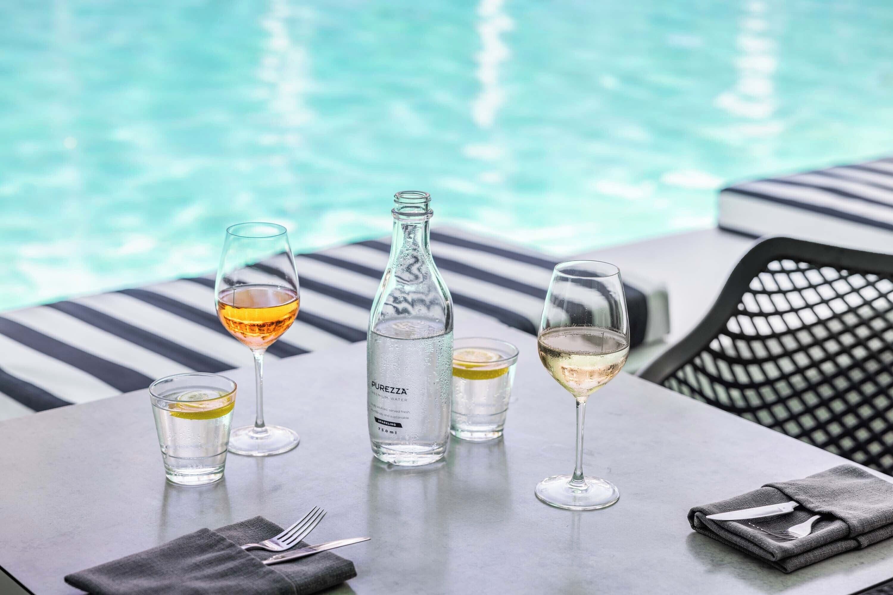 A table with a bottle of water, two glasses of wine, and a glass of water by a poolside.
