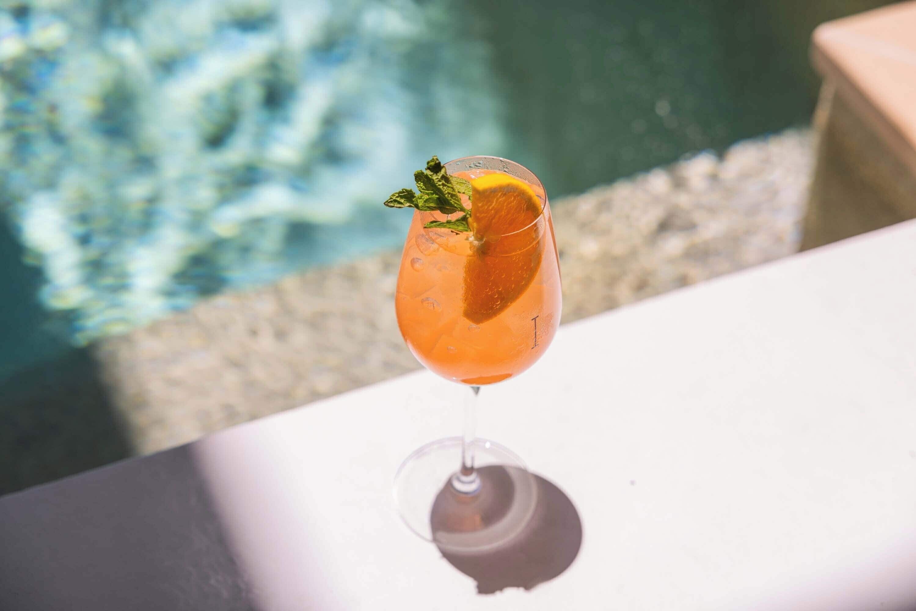 A glass of orange-colored drink with a slice of orange and a mint leaf on a white table near a pool.
