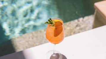 A glass of orange-colored drink with a slice of orange and a mint leaf on a white table near a pool.