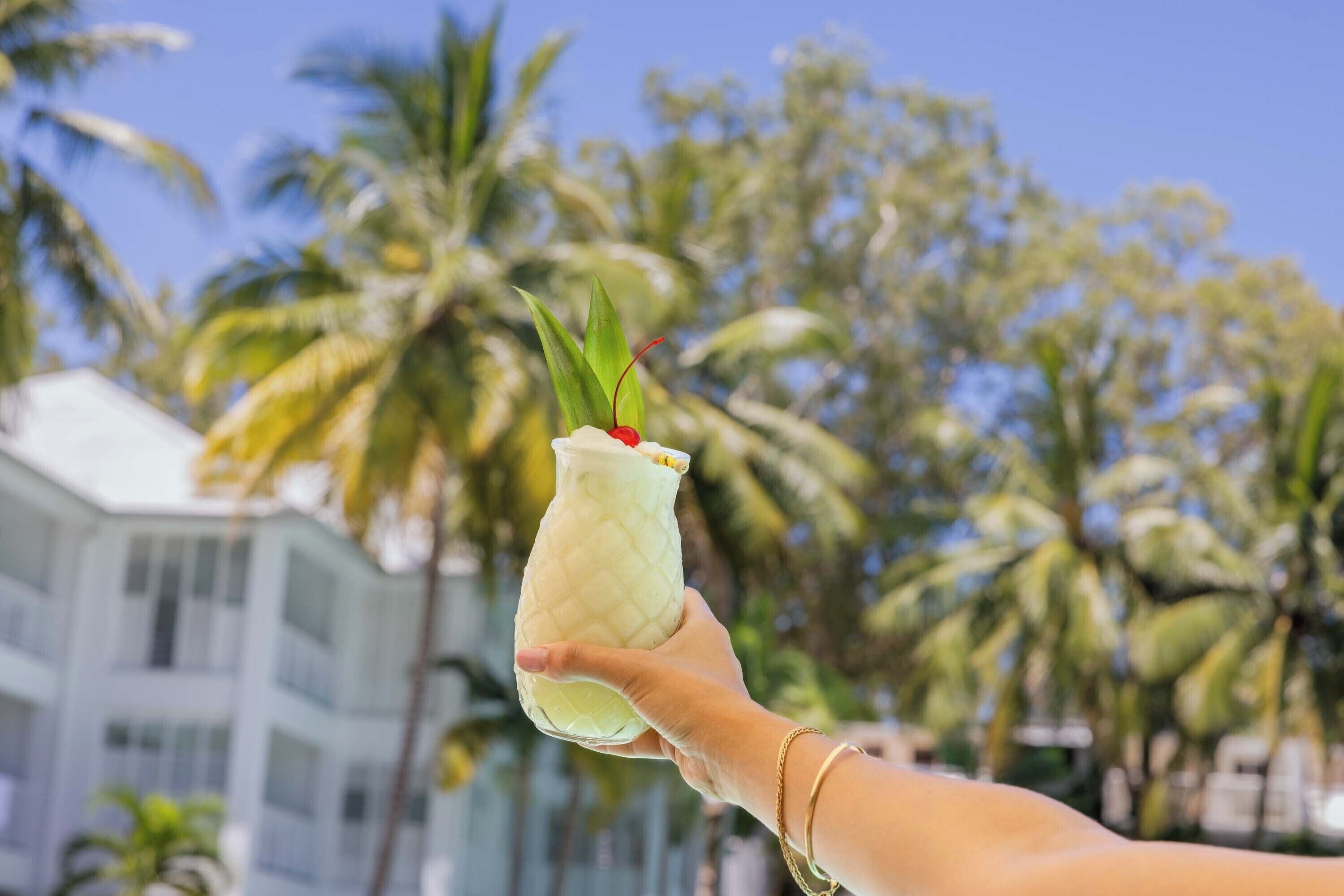 A hand holding a drink with a straw and a cherry, palm trees, and a building in the background.