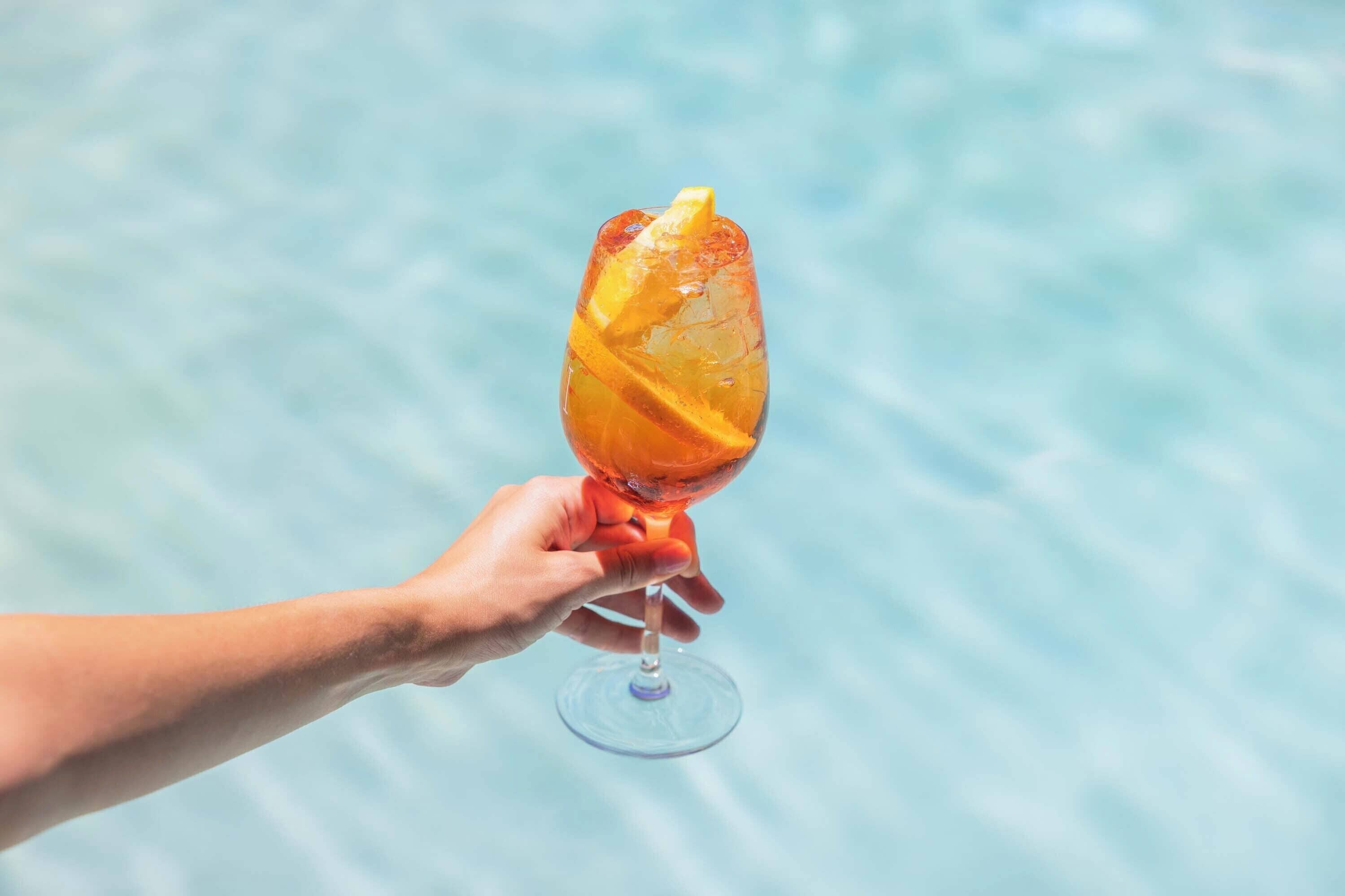 Una mano sosteniendo un vaso con una rodaja de naranja, sobre un cuerpo de agua.