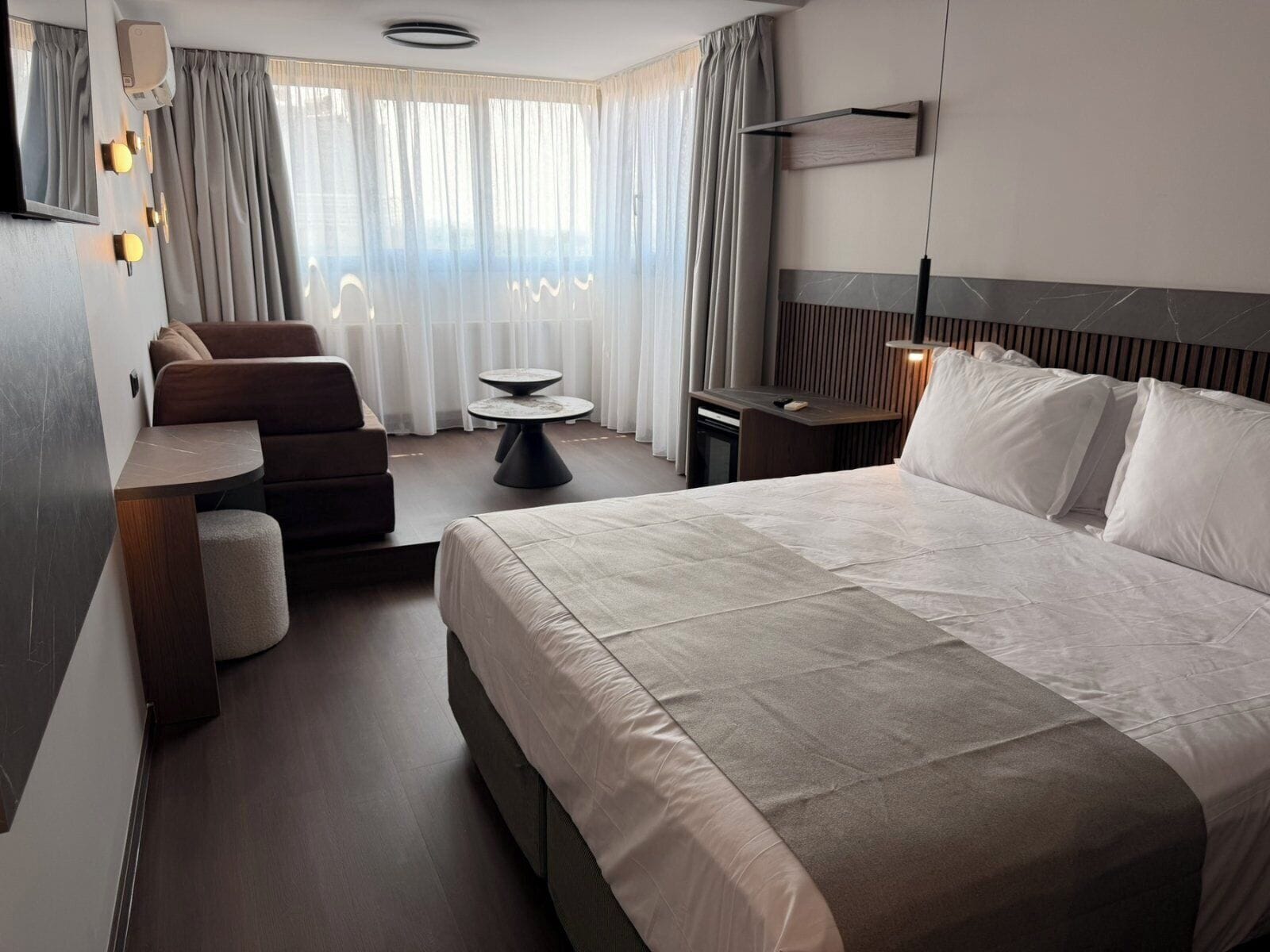 A modern hotel room with a large bed, a sofa, a small table, and a desk.