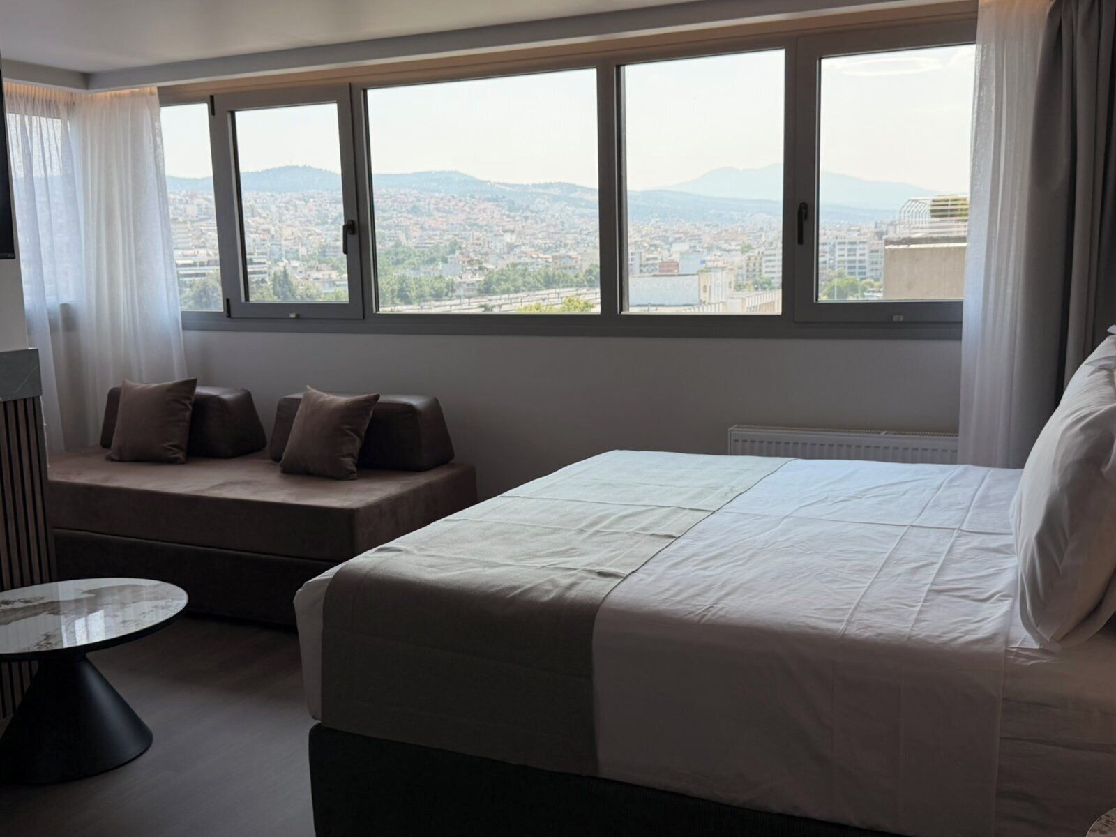 A hotel room with a bed, a seating area with cushions, a round table, and a large window with a city view.