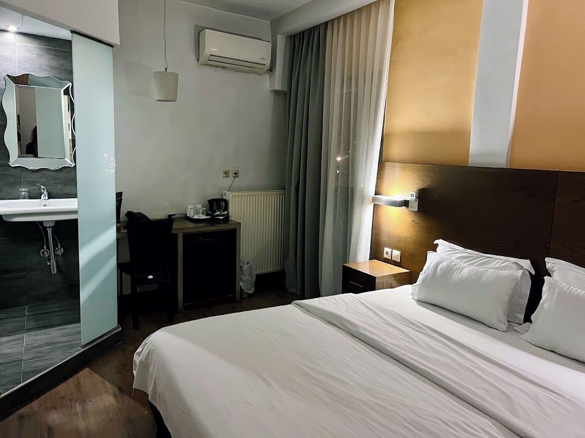 A hotel room with a bed, a desk, a chair, a mirror, a sink, and a window with curtains.