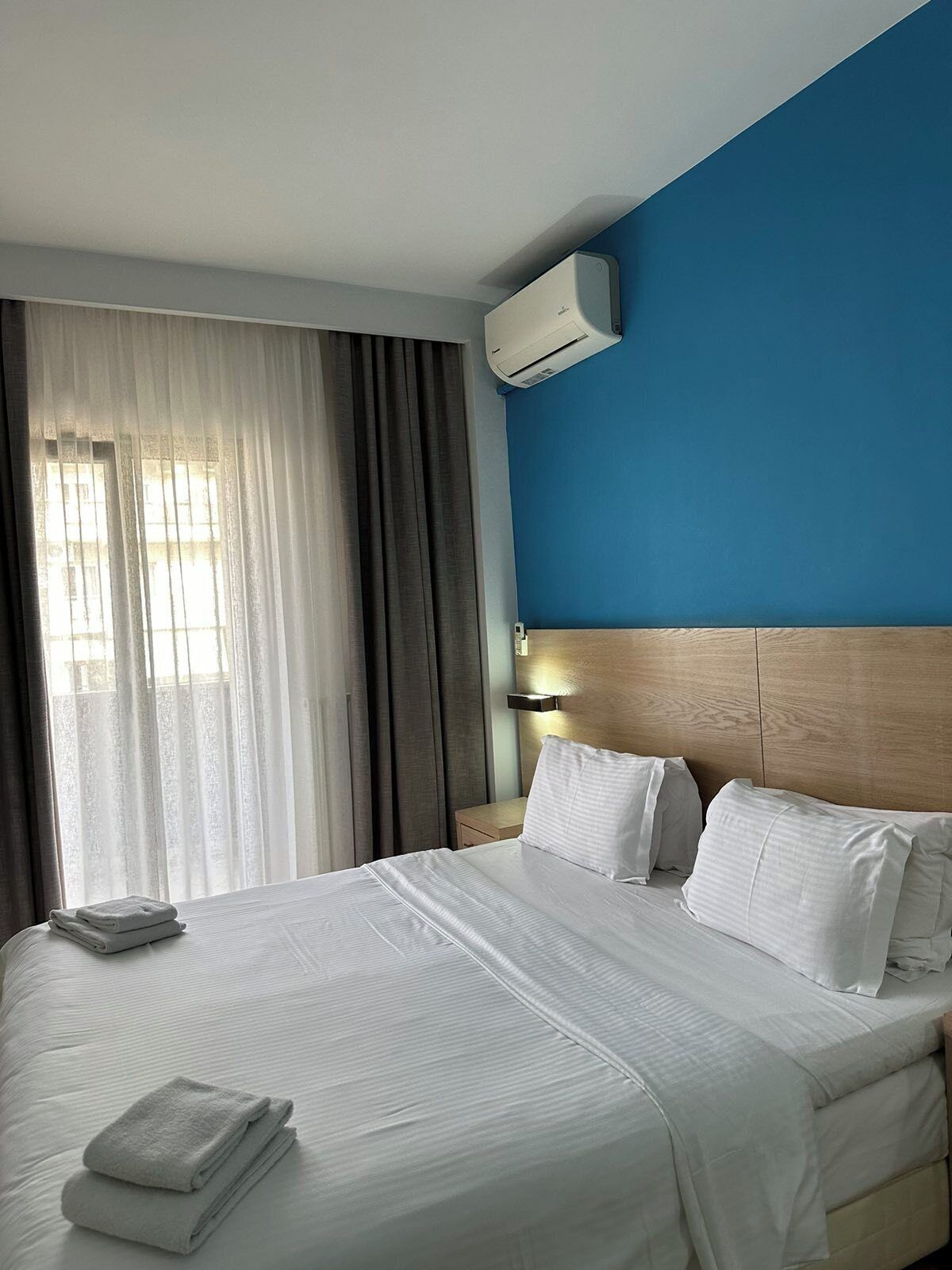 A hotel room with a bed, curtains, an air conditioning unit, and a nightstand.