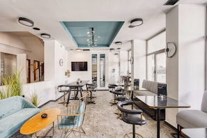 A modern interior with a long dining table, bar stools, a sofa, and a large window providing natural light. - Hotel Rotonda (Thessaloniki)