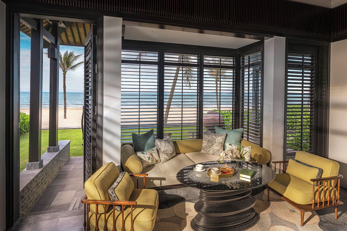 A modern living room with a view of the beach, featuring a sofa, chairs, a coffee table, and large windows with blinds.