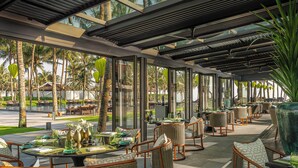 An outdoor dining area with wicker furniture, glass tables, and large windows overlooking a pool and palm trees. - Four Seasons Resort The Nam Hai, Hoi An, Vietnam (Da Nang)