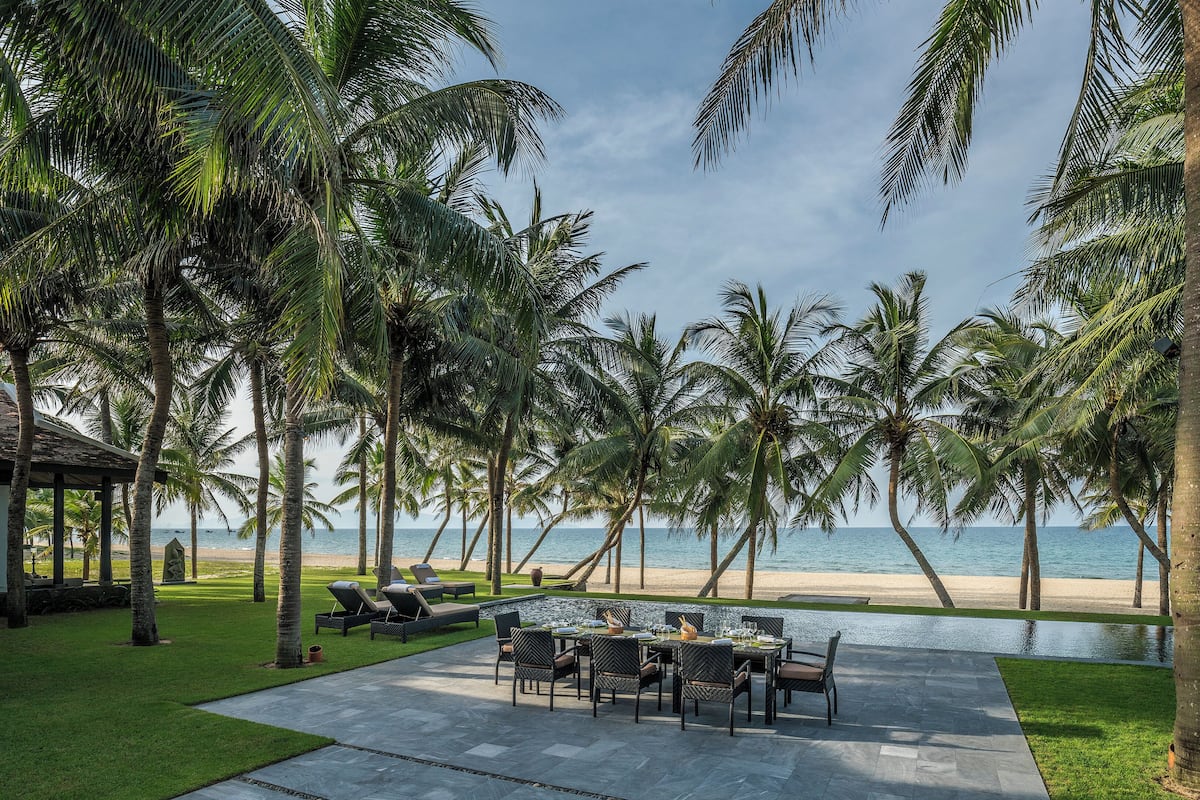 A poolside dining area with a view of the beach and ocean, surrounded by palm trees.