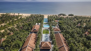 Luchtbeeld van een resort met meerdere bungalows, een centraal gelegen zwembad en een strand op de achtergrond.