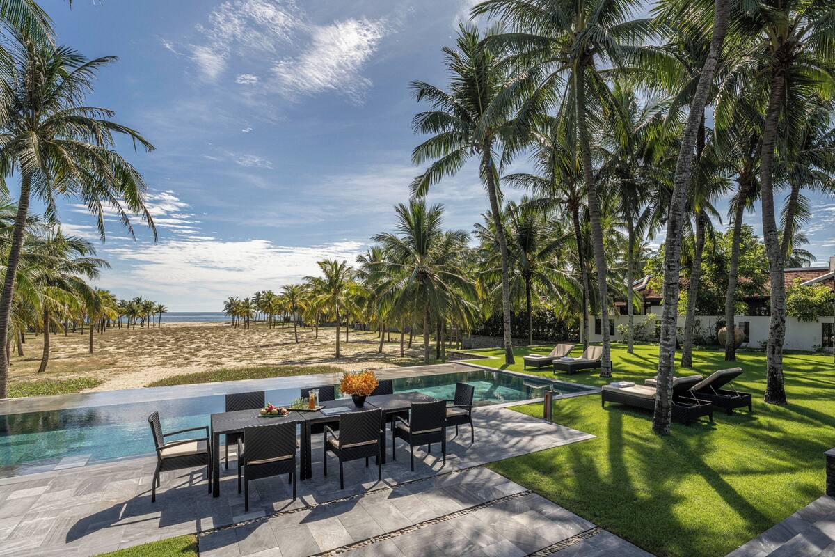 A poolside area with a dining table and chairs, surrounded by palm trees and a view of the beach.