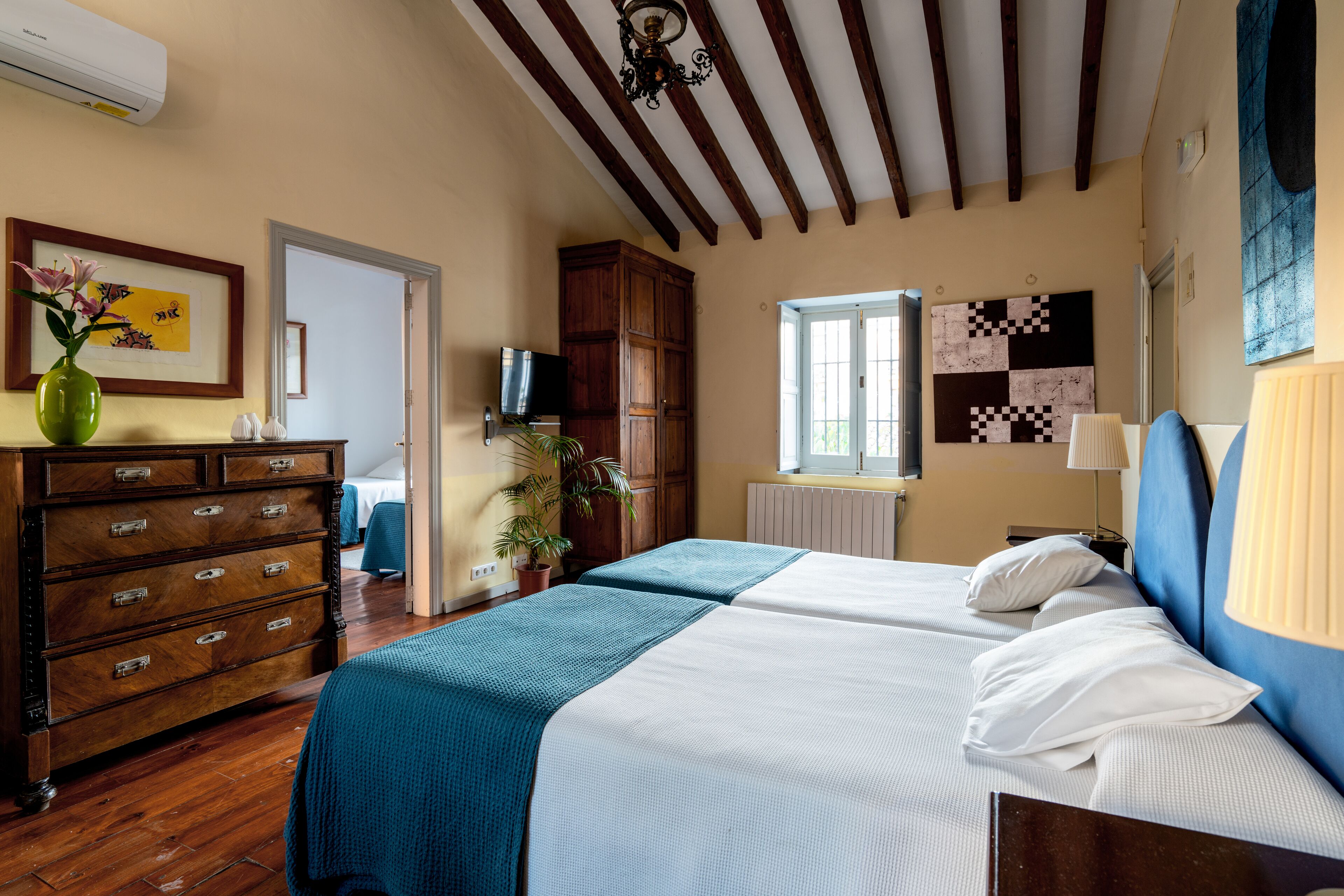 A hotel room with two beds, wooden furniture, a TV, a window with a view, and a wall with a quilt pattern.