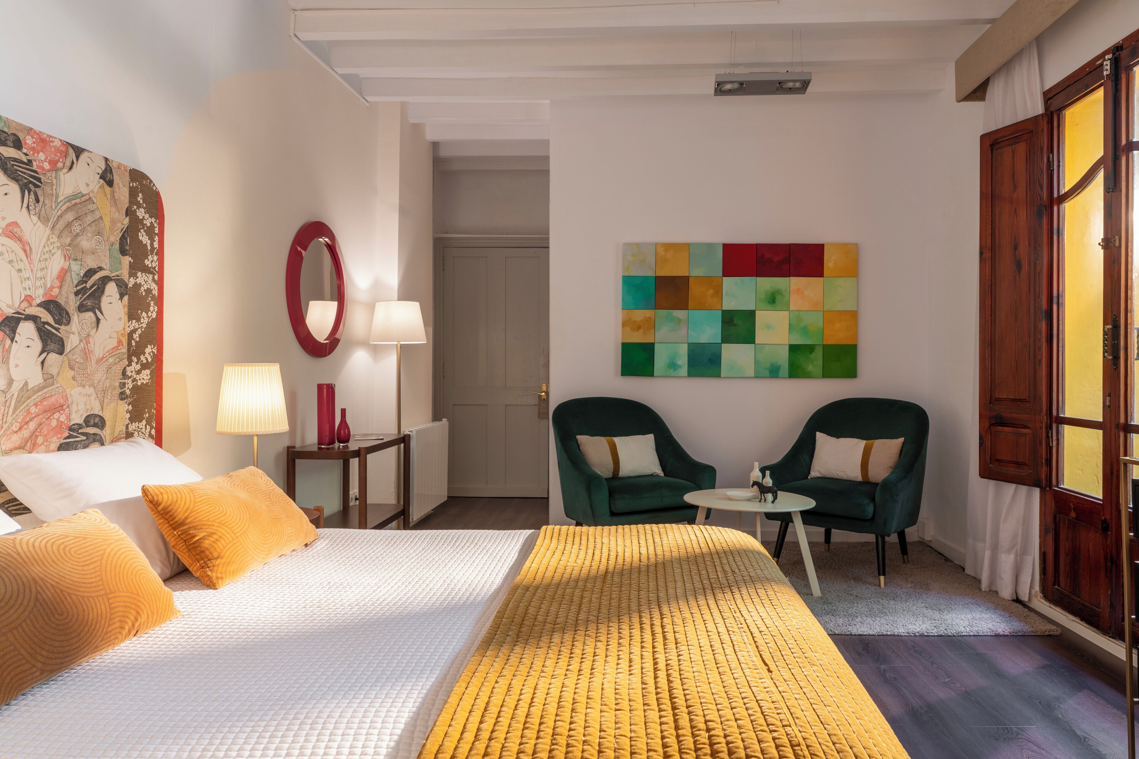 A hotel room with a bed, two chairs, a small table, a lamp, a mirror, and a colorful artwork on the wall.