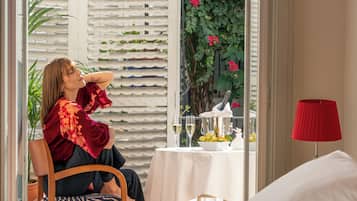 Une chambre avec un lit, une table surmontée d’une lampe rouge, et une vue sur un jardin où poussent des fleurs roses.
