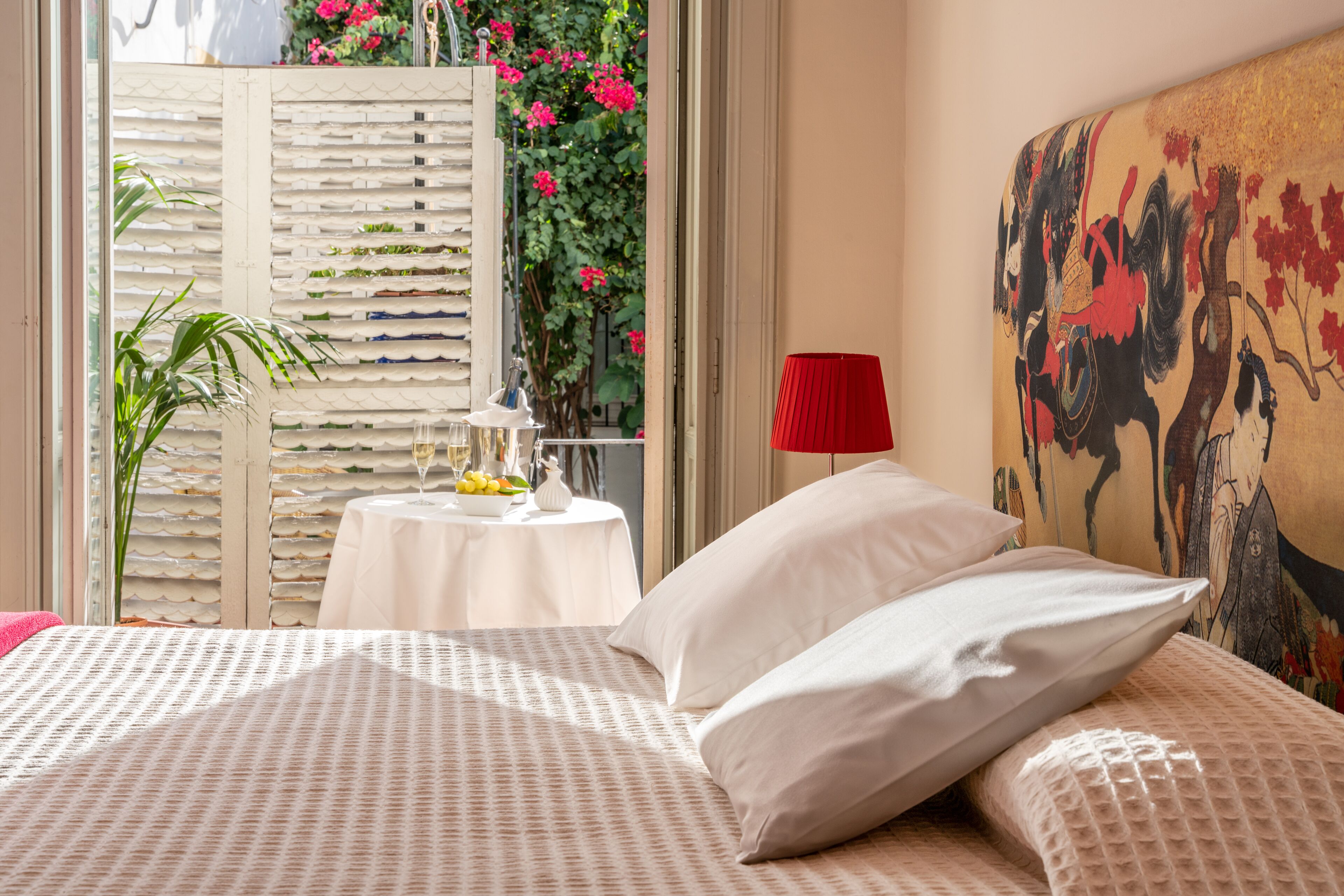 Un dormitorio con cama, almohadas, una mesa con un florero y frutas, una lámpara roja y un cuadro en la pared.