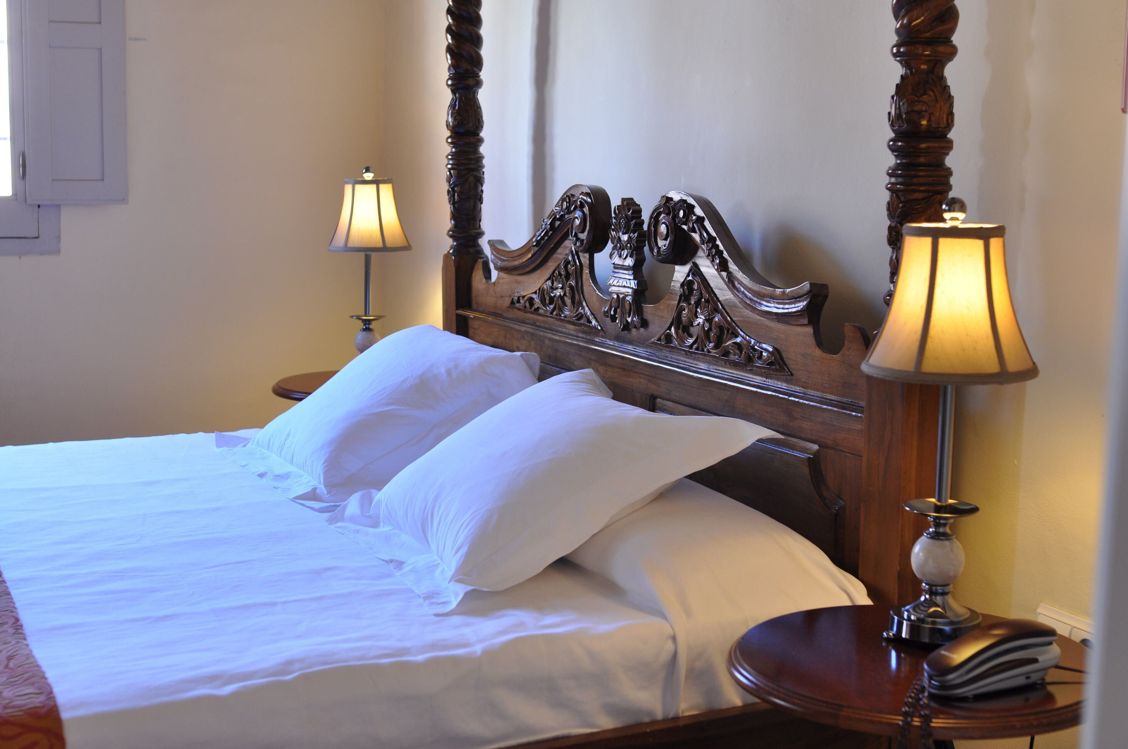 Una cama bien hecha con sábanas y almohadones blancos, un cabecero de madera con intrincados grabados, y una mesita de noche con una lámpara y un libro.