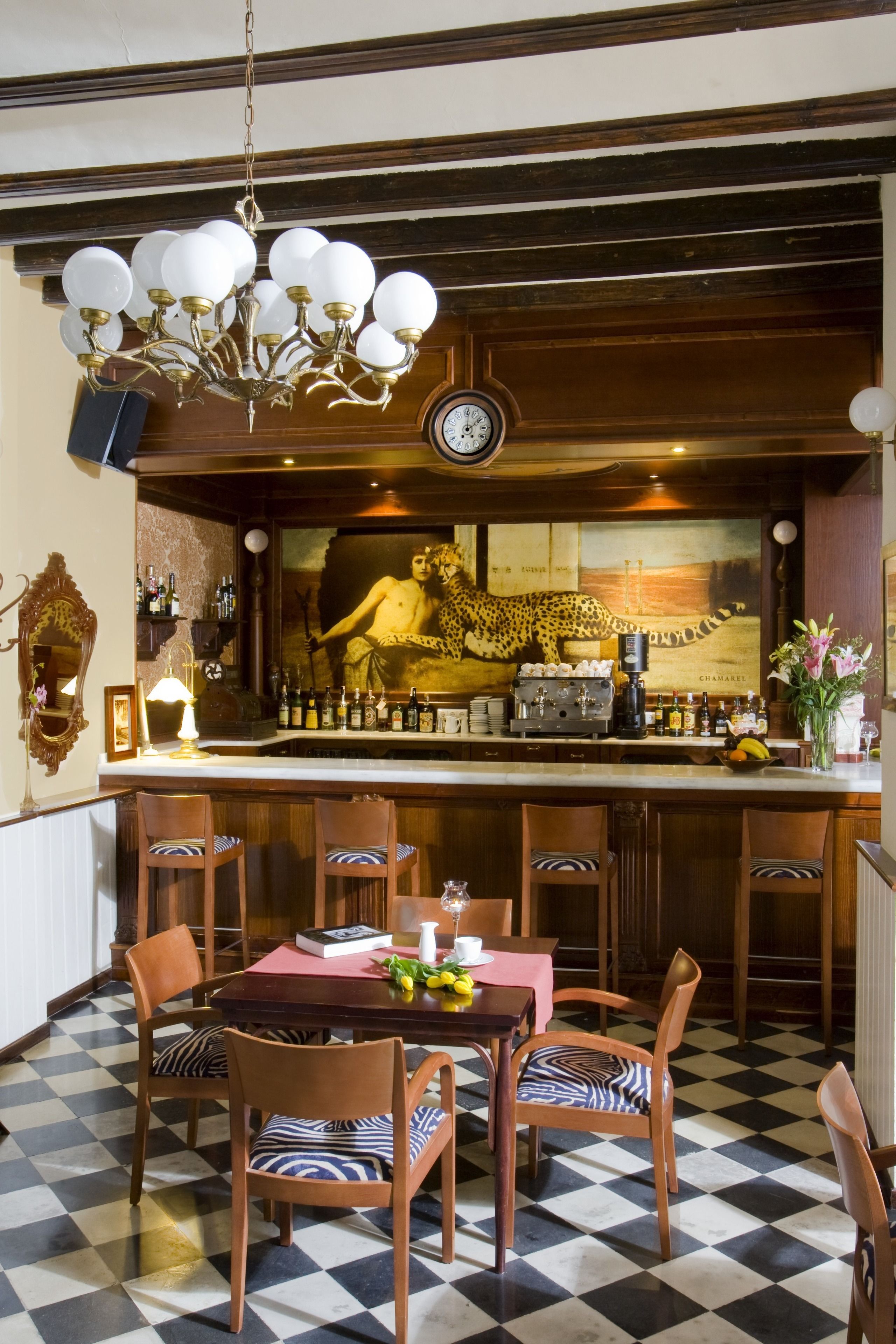 A bar area with a chandelier, a clock, and a leopard mural. There are bar stools and a table with chairs.