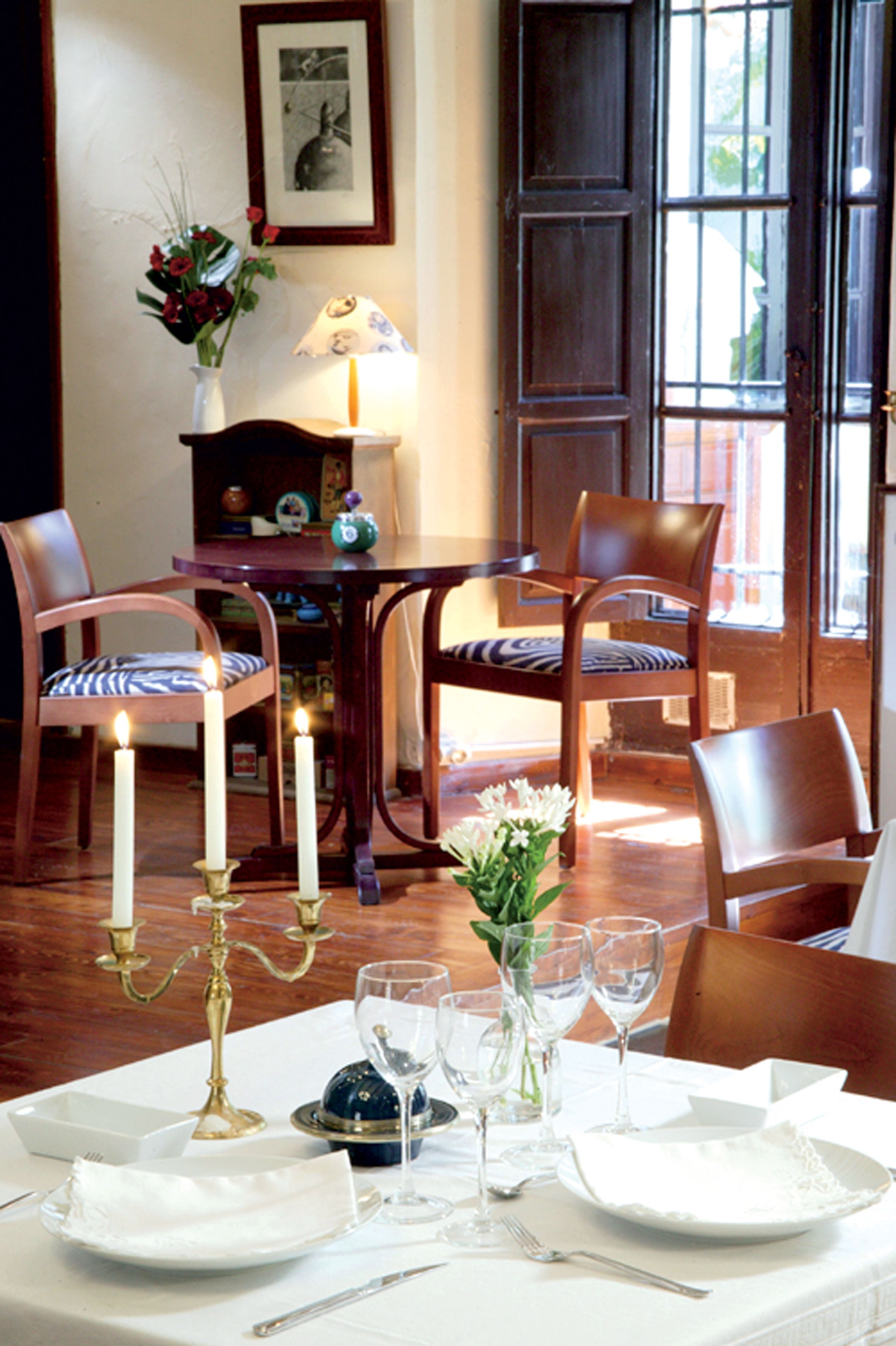 Comedor con una mesa dispuesta para cuatro, con platos blancos, cubiertos y un candelabro. Hay dos sillas con cojines a rayas azules y blancas, y un aparador de madera con adornos.