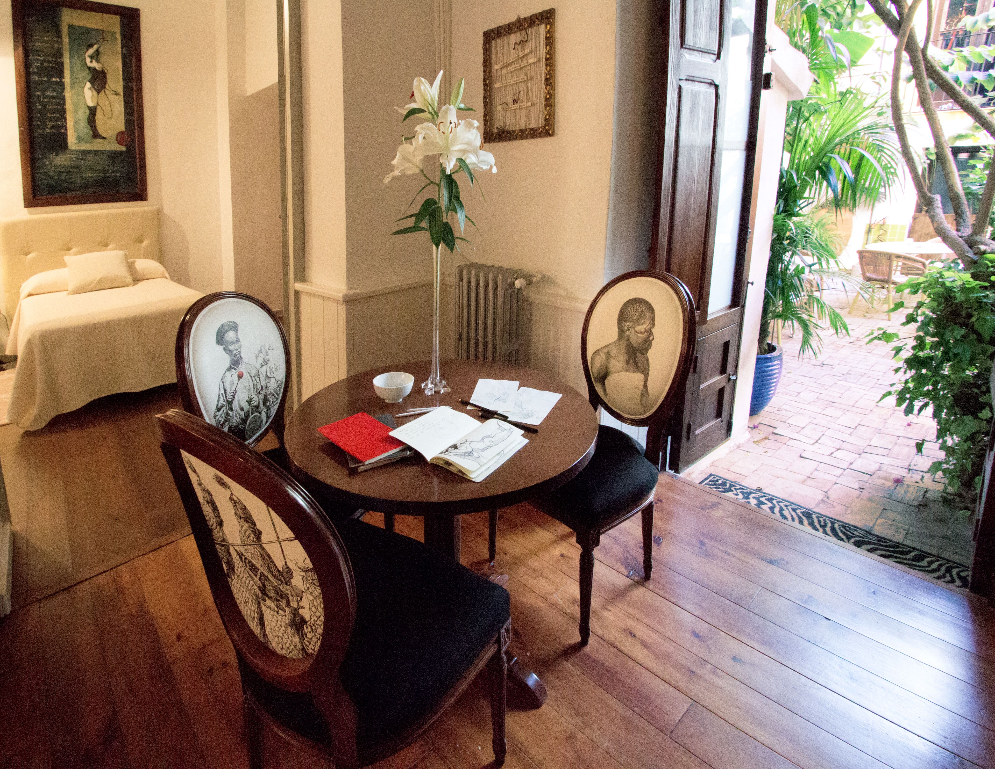 Un comedor con una mesa redonda, sillas y un jarrón de flores. Hay cuadros en las paredes y una cama al fondo.