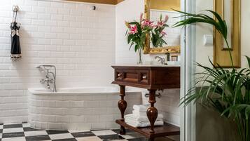 Un baño con pared de azulejos blancos, suelo a cuadros blanco y negro, tocador de madera, bañera blanca y espejo con marco dorado.
