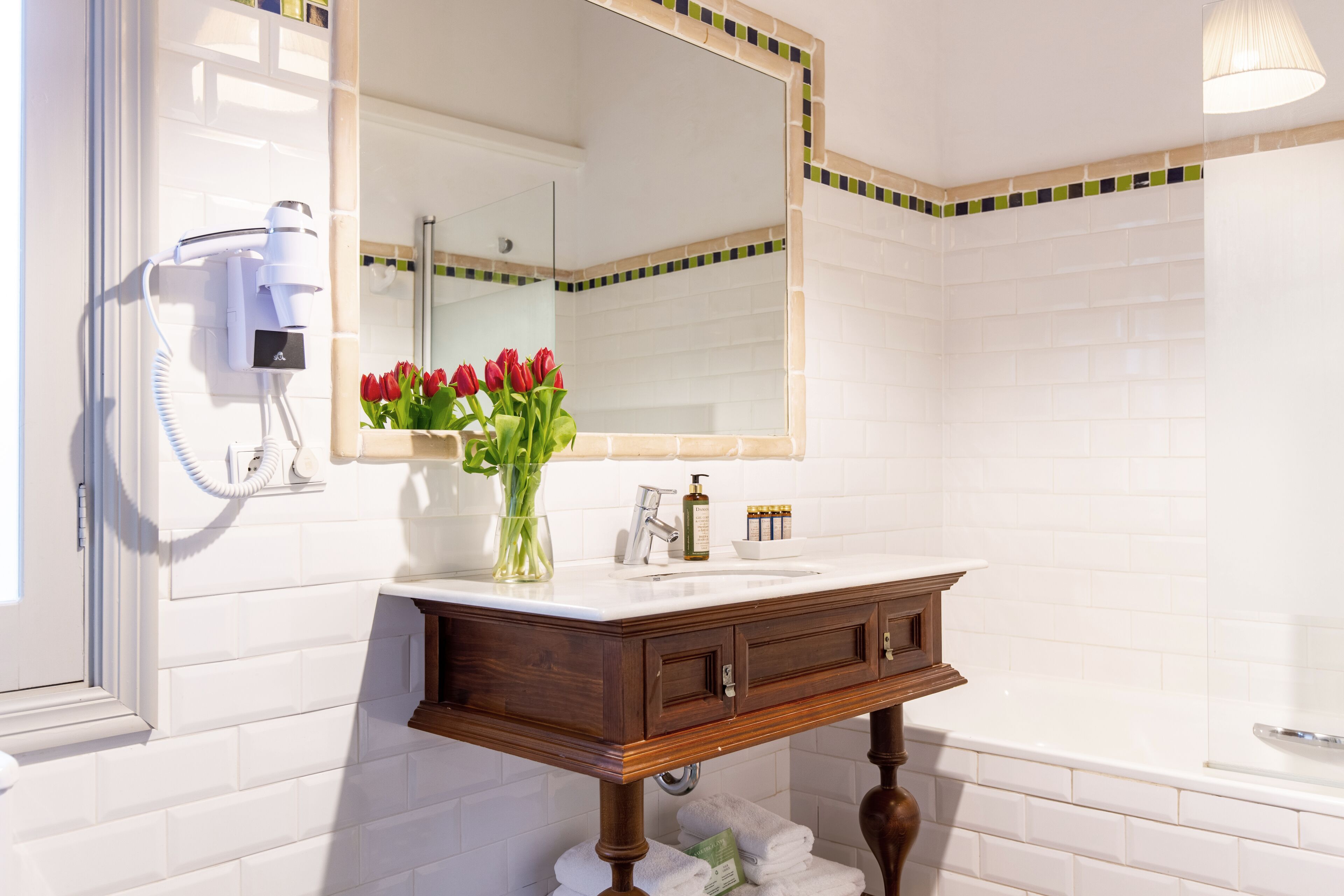 Un baño con pared de azulejos blancos, tocador de madera con espejo, secador de pelo y un florero con flores rojas.