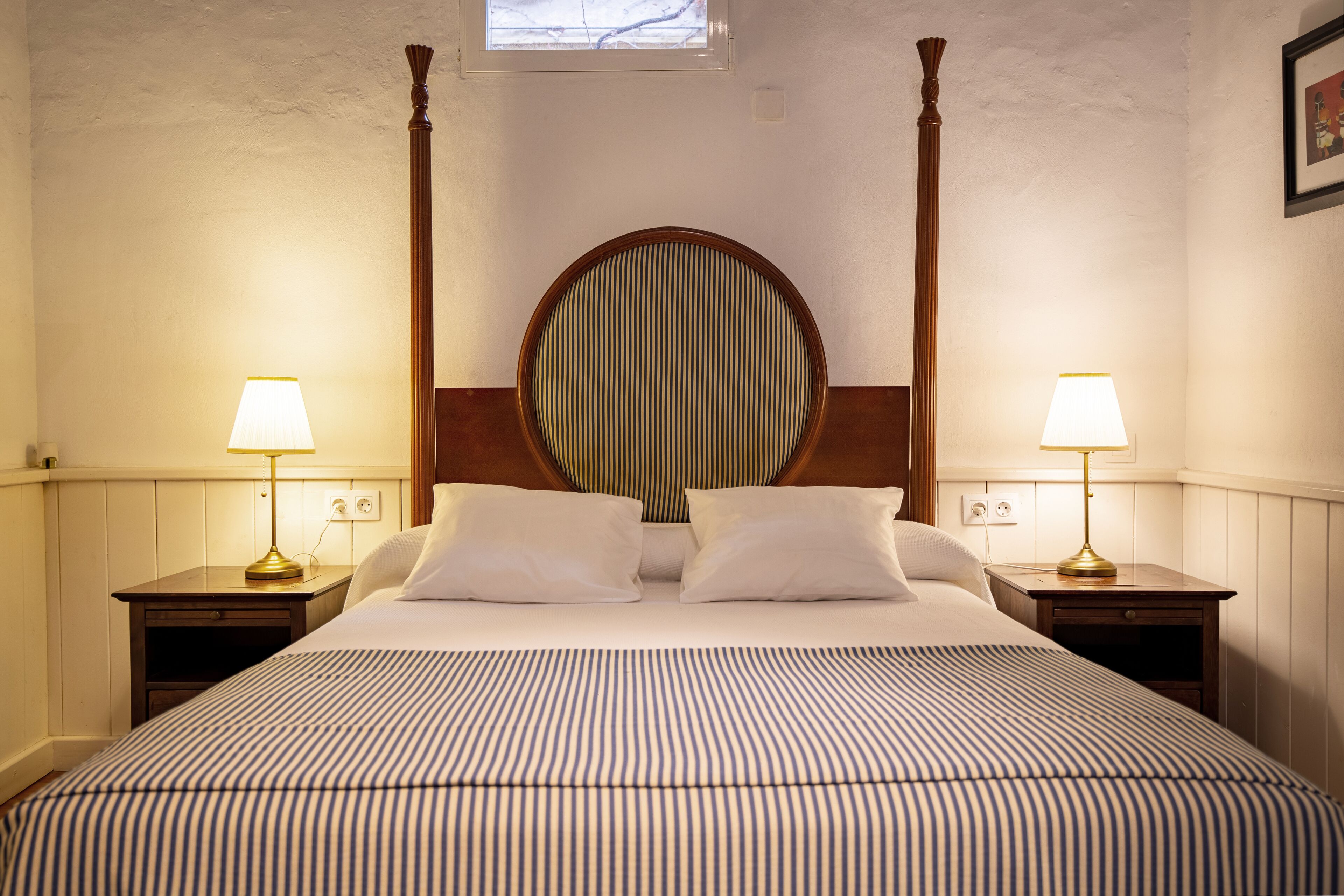 A four-poster bed with a round mirror above it, flanked by two bedside tables with lamps, in a room with white walls and wooden trim.