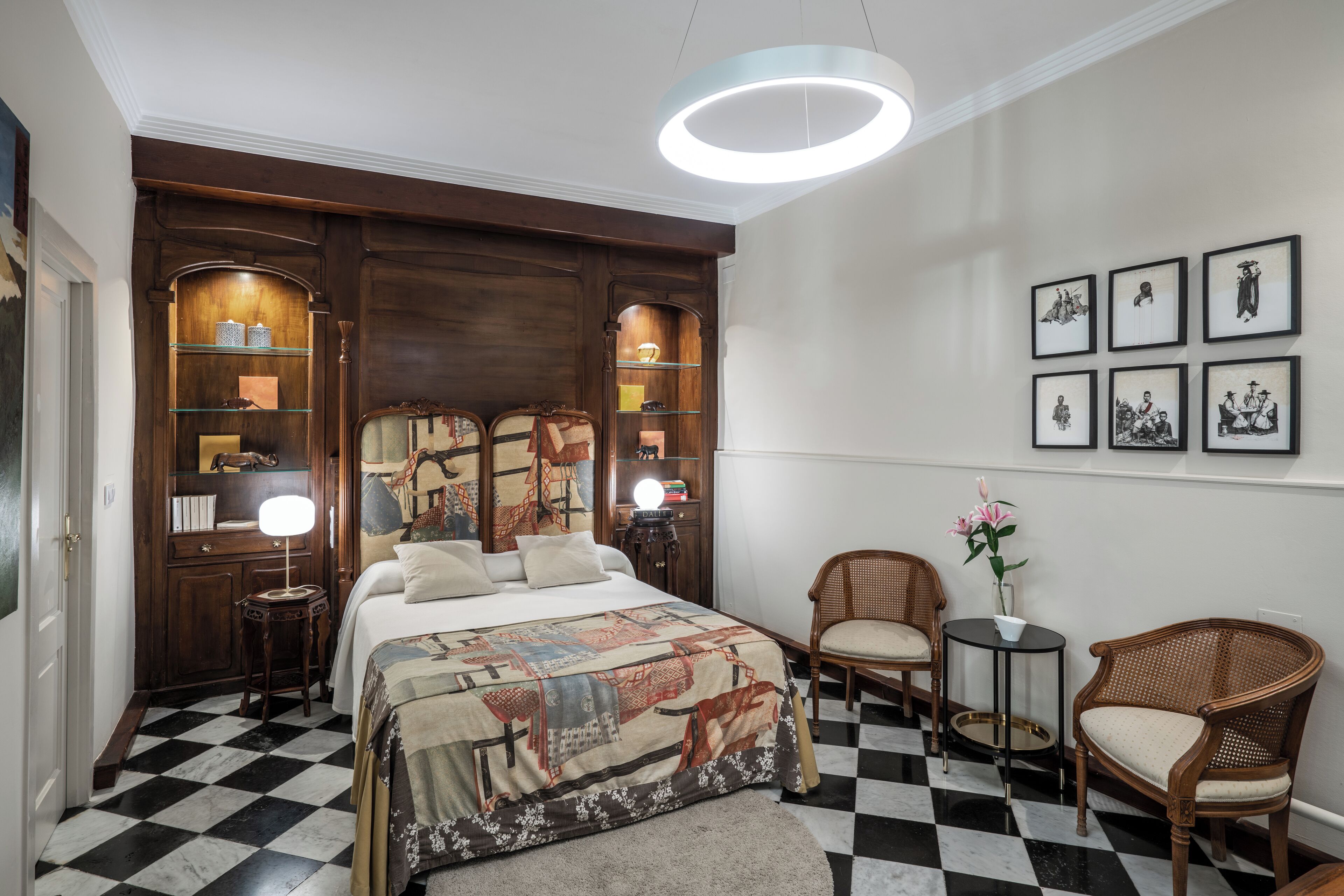 Un dormitorio con suelo a cuadros, una cama grande con edredón estampado, cabecero de madera, mesitas de noche con lámparas, una pequeña mesa redonda con una silla y cuadros en la pared.