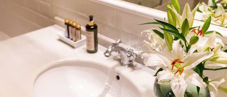 A bathroom sink with a mirror above it, a bottle of shampoo, and a vase with white flowers.