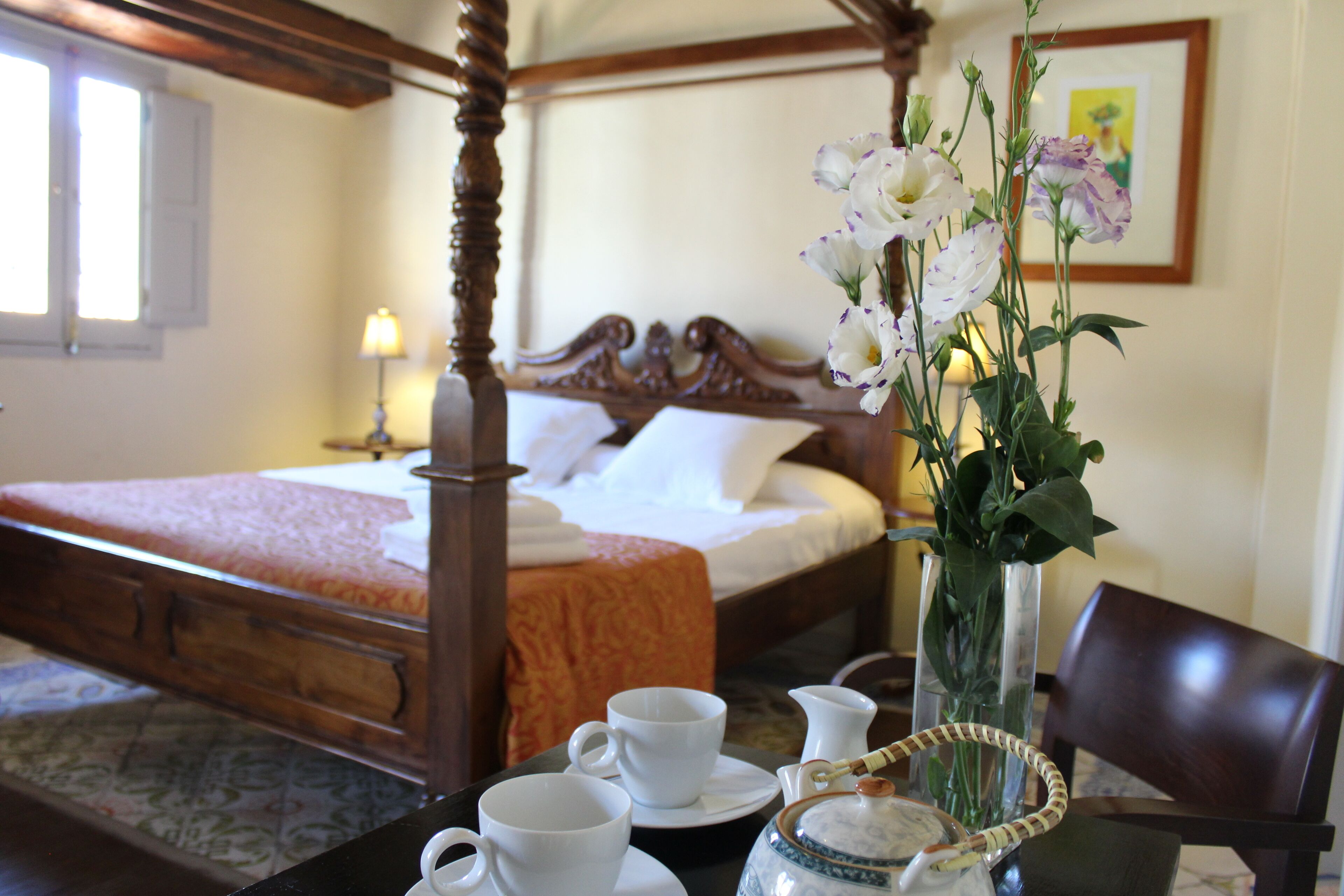 A hotel room with a four-poster bed, a vase with flowers, and a table with cups and a pitcher.